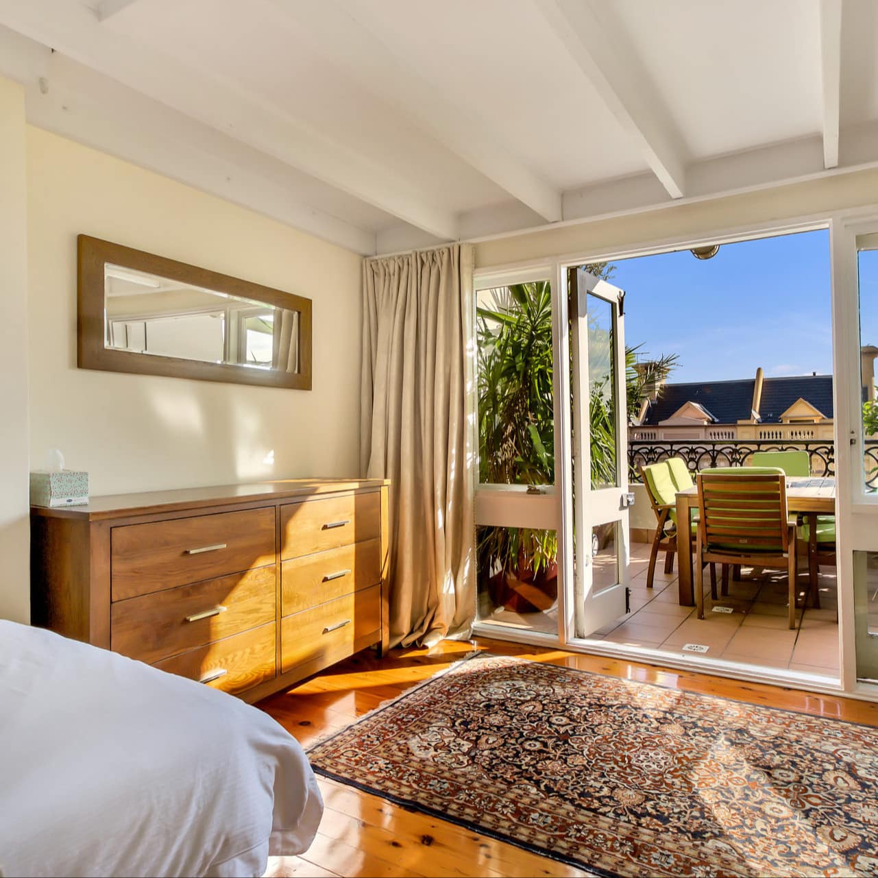 Sunny bedroom with hardwood floors, a queen size bed, persian rug, and attached balcony with city views.
