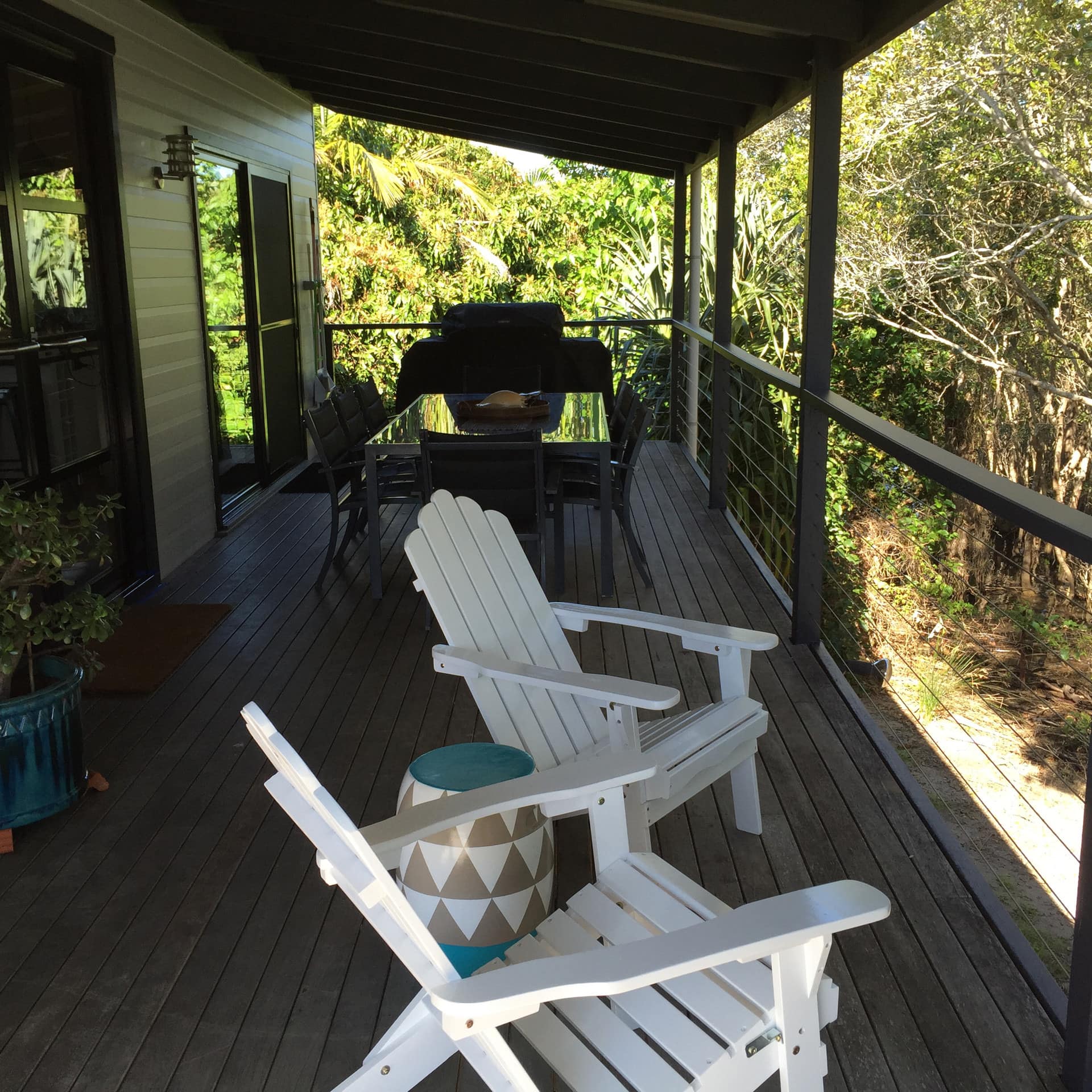 Covered veranda surrounded by thick vegetation, wood floors, white Adirondak chairs and glass dining set.