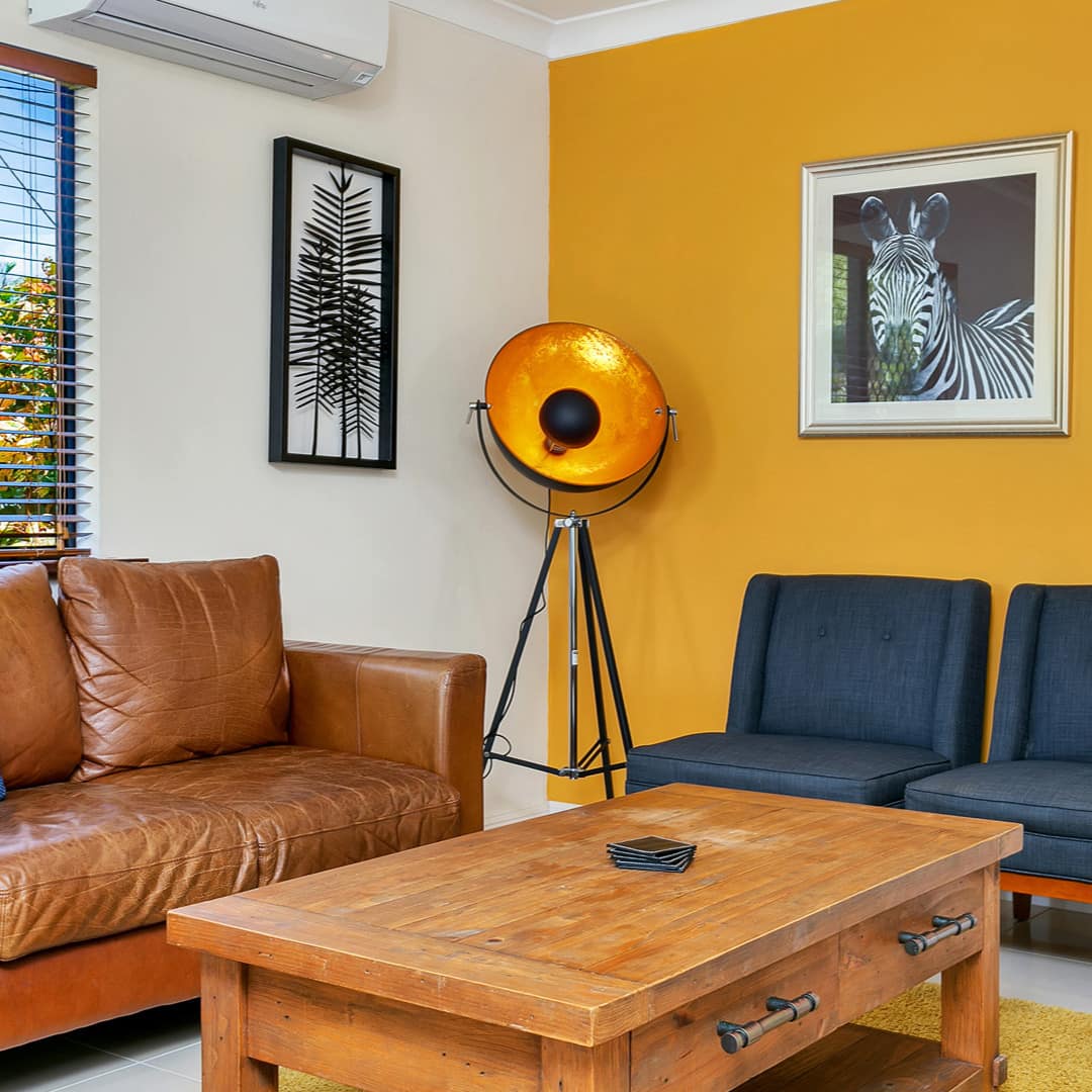 Modern living room with yellow accent wall, brown leather sofa, navy blue chairs, wooden coffee table, black and white art.