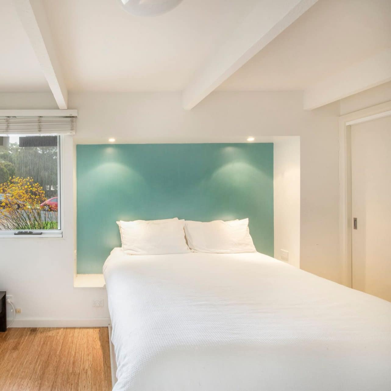 Modern bedroom with hardwood floors, white walls, bed set in recessed wall with teal backsplash, and floral art.