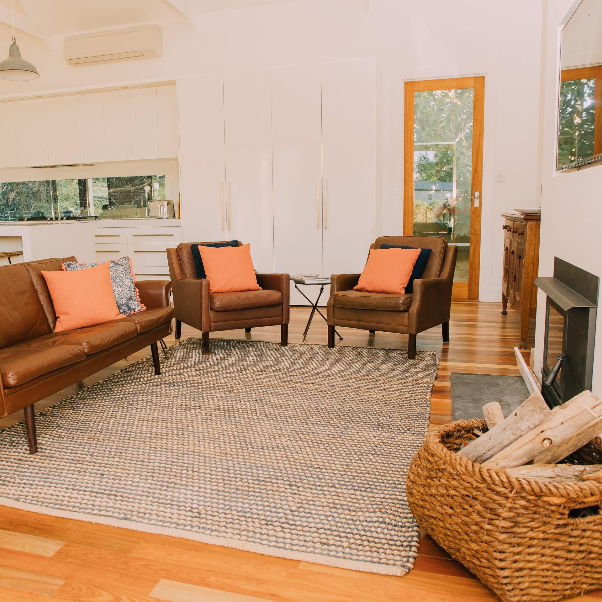 Open living room and kitchen, matching brown leather sofa and chairs, fireplace, wicker basket with chopped wood, woven rug.
