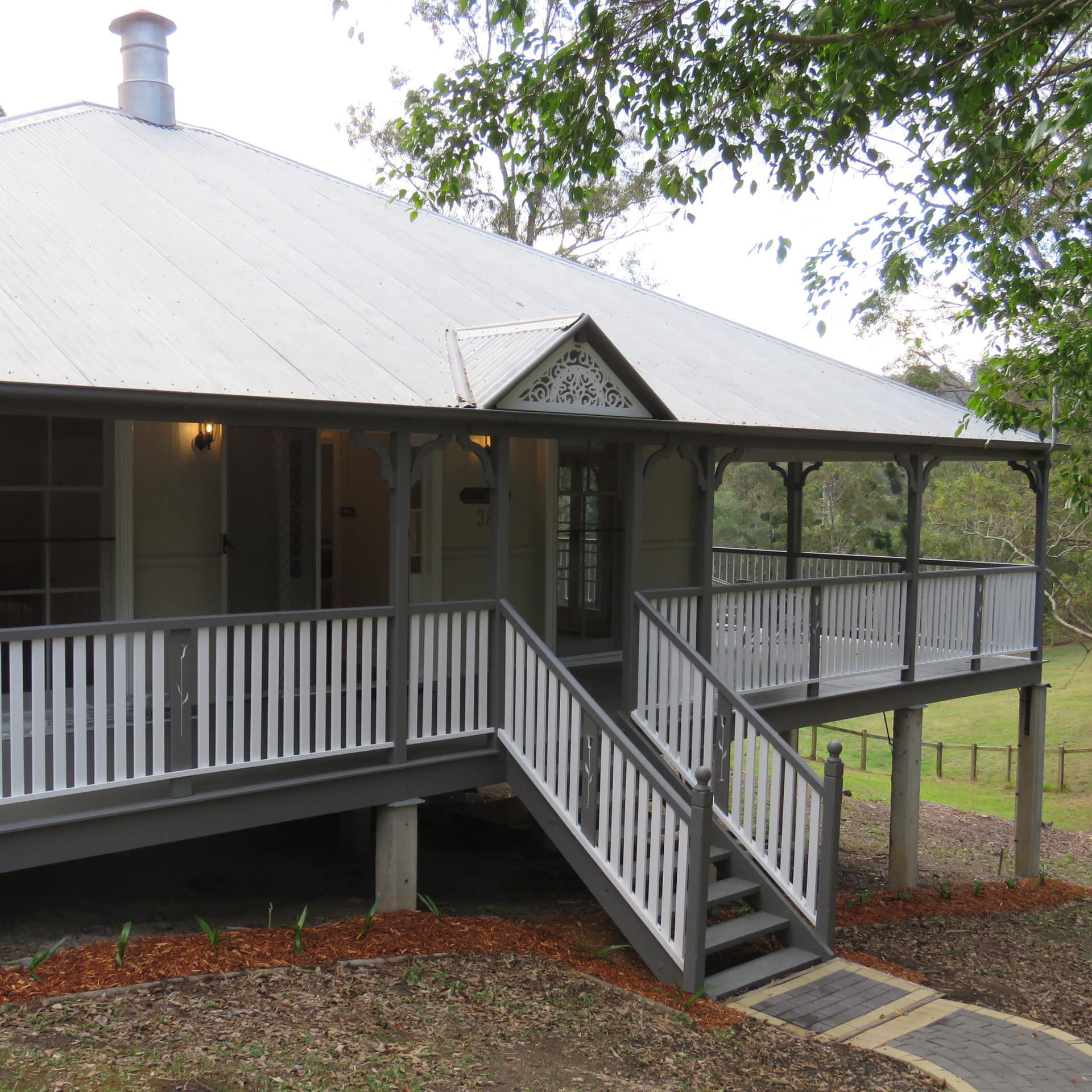 1-storey grey holiday cottage with wrap-around porch and metal roof set amidst farmland and backed by trees