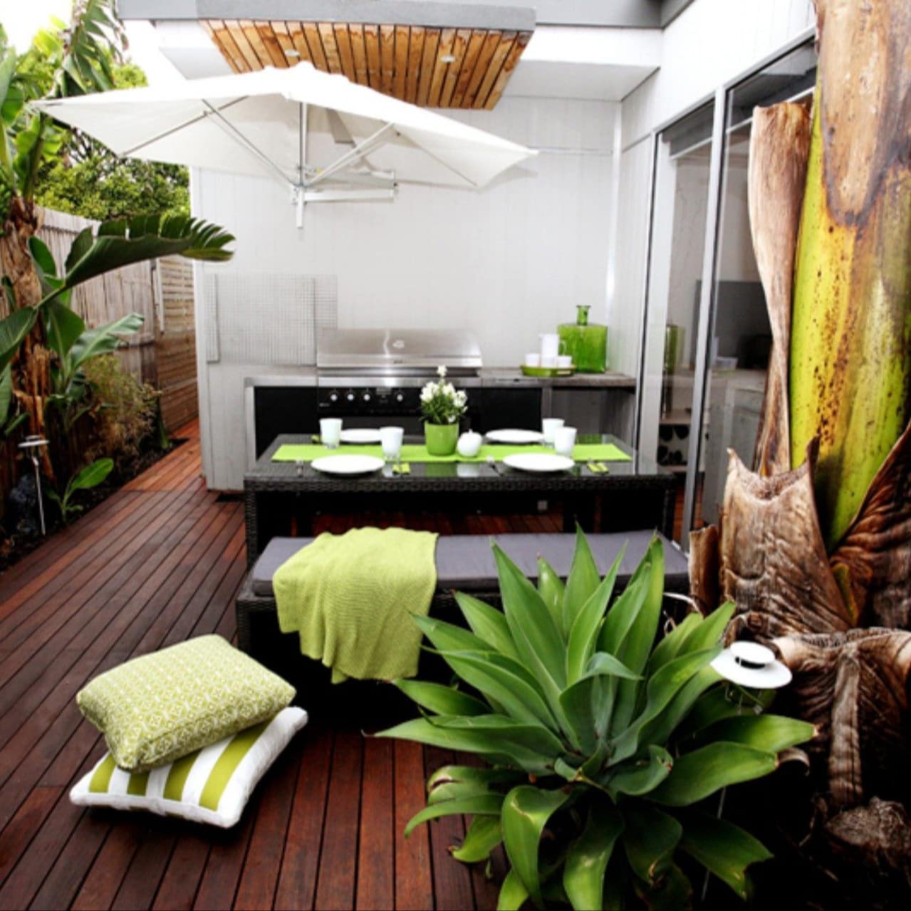 Covered wooden deck with black dining set, grill, sliding glass doors, decorated with lush tropical plants.