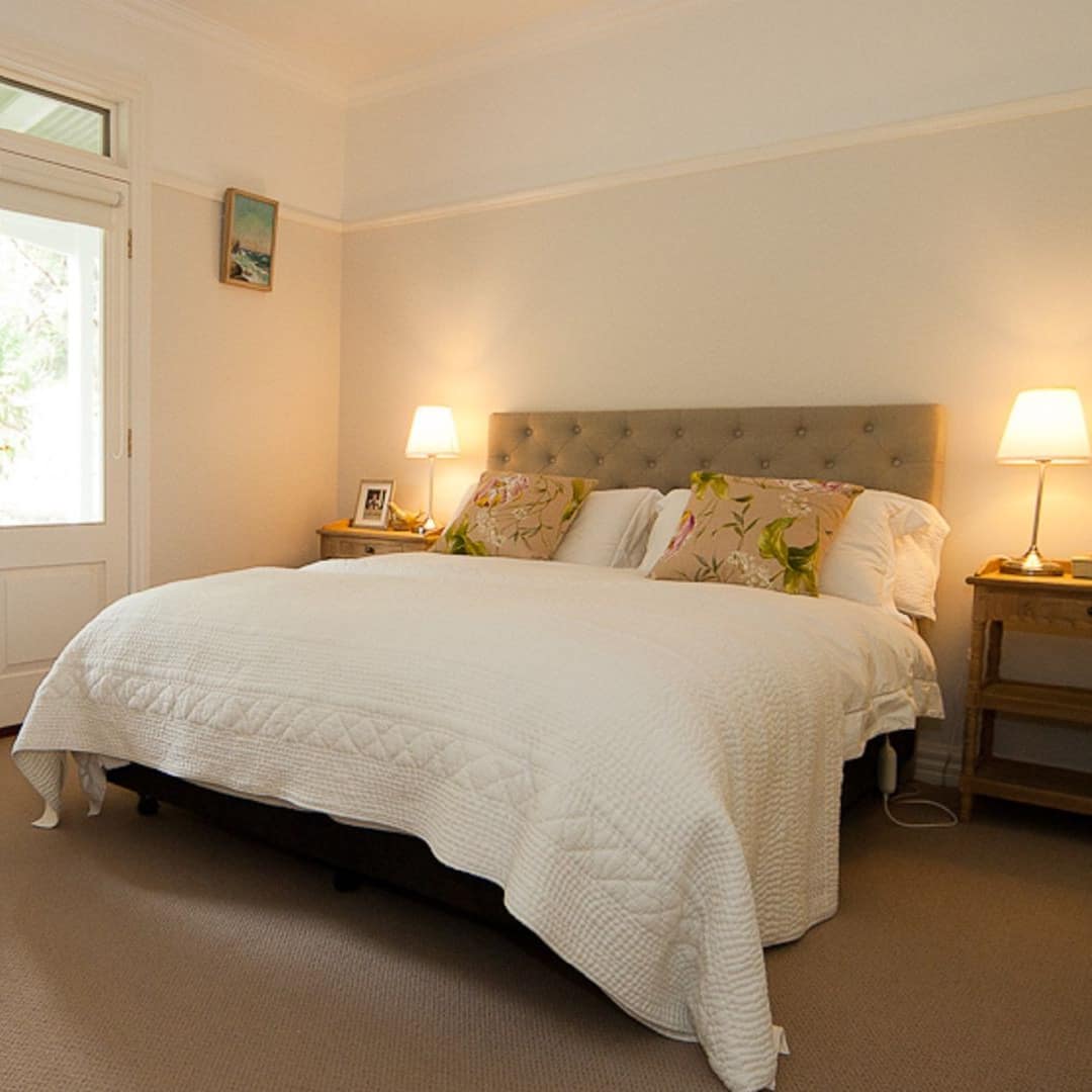 Spacious bedroom with tan carpet, white walls, king size bed, white bedding and floral pillows, glass double doors.