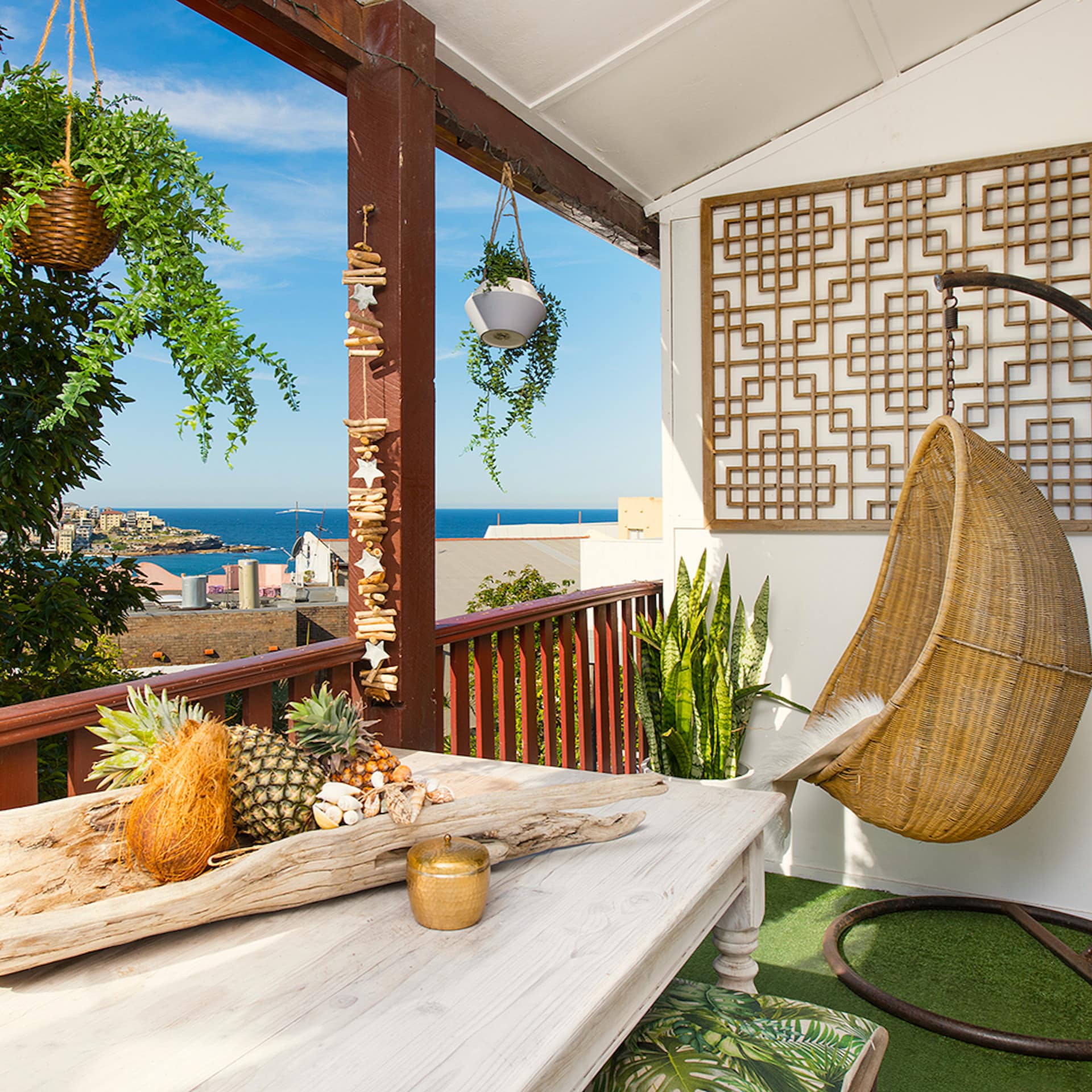 A covered porch with a hanging rattan chair, and wooden table with fresh fruit on it, overlooking the beach.