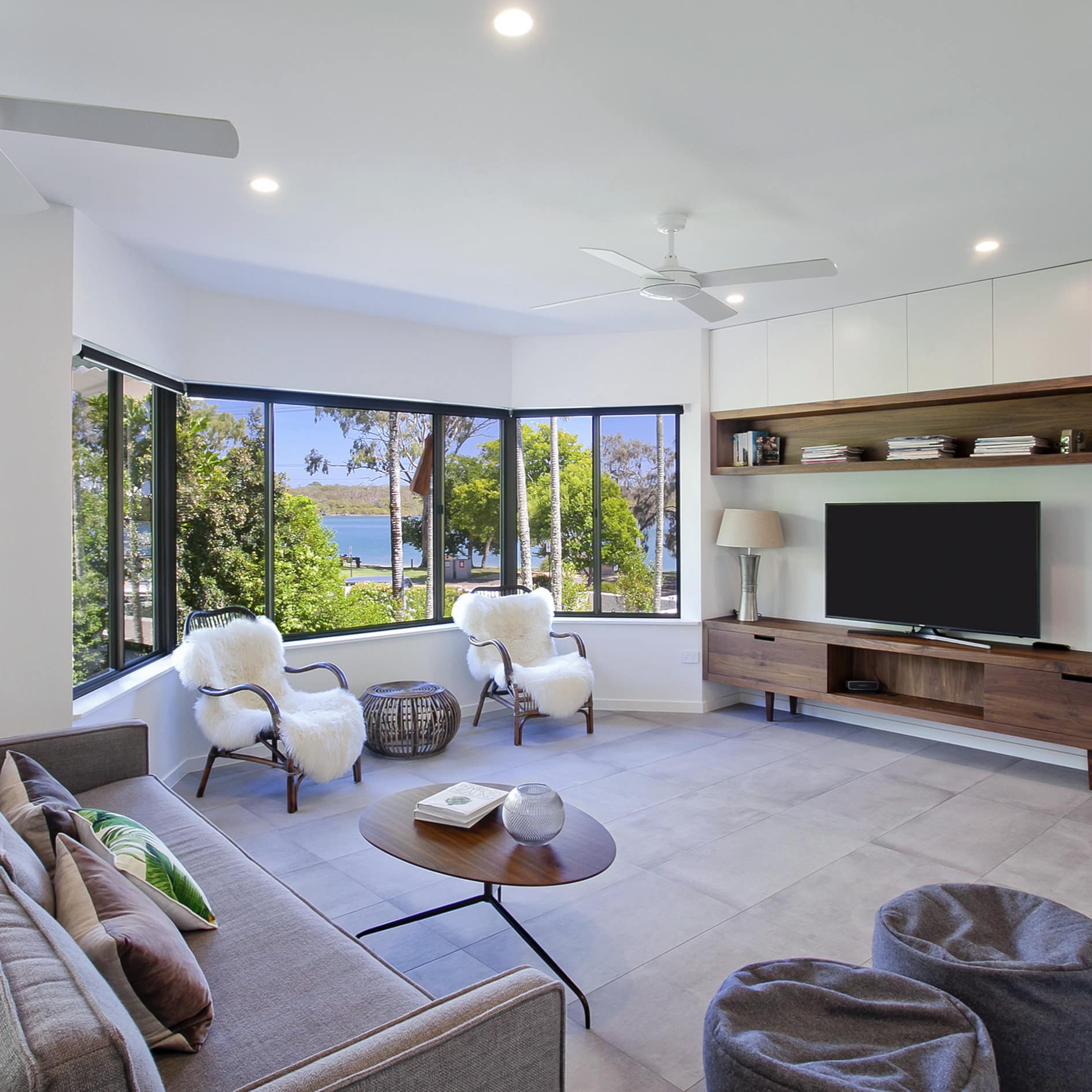 Modern living room, grey sofa, black bamboo chairs with sheepskin covers, flatscreen TV, wide bay windows with ocean views.
