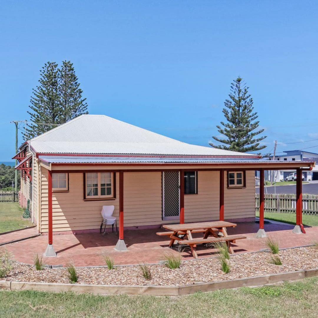 Front lawn of a white cottage with red trim, covered deck, picnic table, raised garden beds, and wood picket fence.