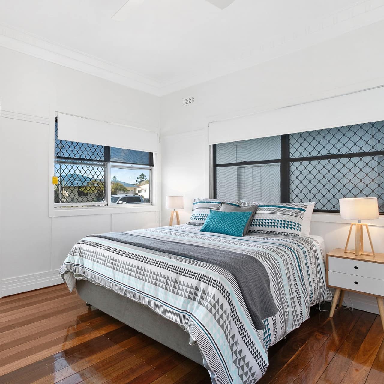 A spacious bedroom decorated in blue and grey with a queen size bed, hardwood floors, and windows with neighborhood views.