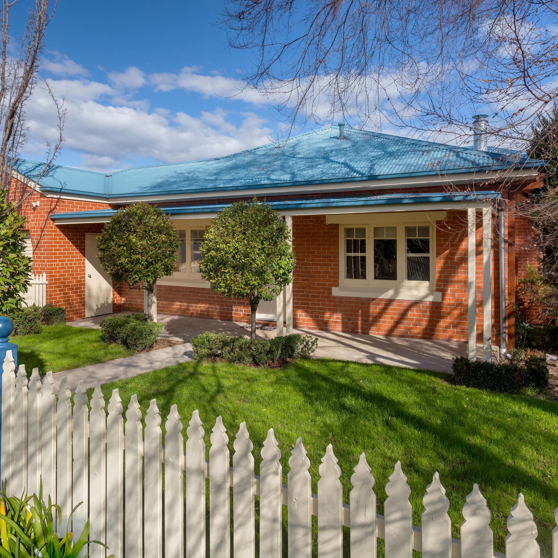 Lovely red brick ranch home with a blue metal roof, a front porch, a lush green lawn and a white picket fence.