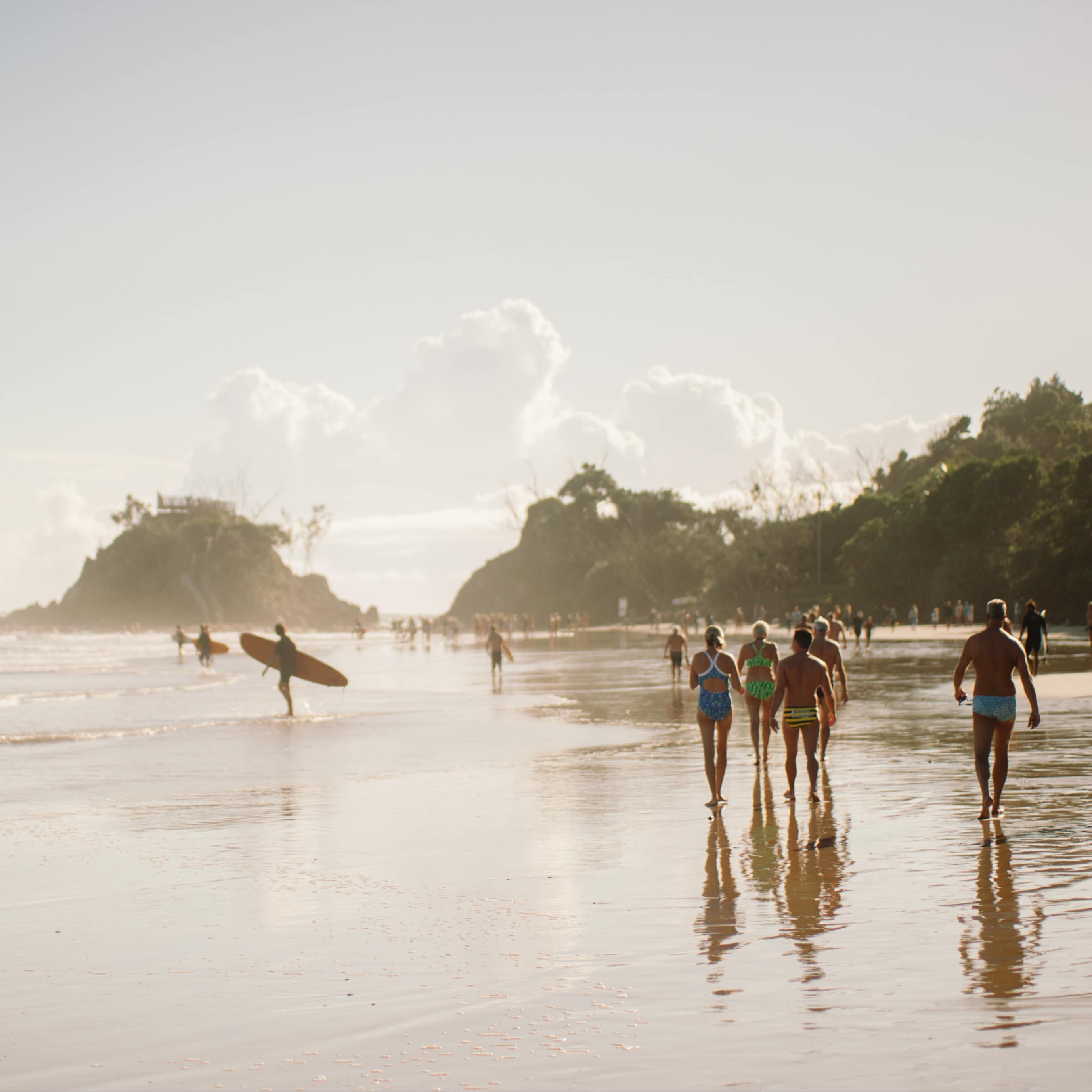 Byron Bay beach with adults walking along the shore, surfers heading out to catch waves and tree-covered hills