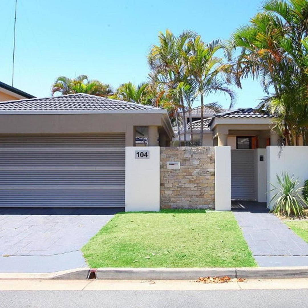View of a cottage with a garage and enclosed by a white and stone wall, surrounded by palm trees.