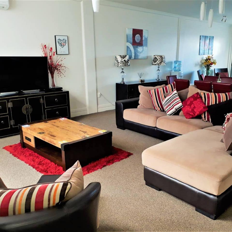 Open living and dining room with beige carpet, tan sectional, wooden coffee table, flatscreen TV, red and black decor.