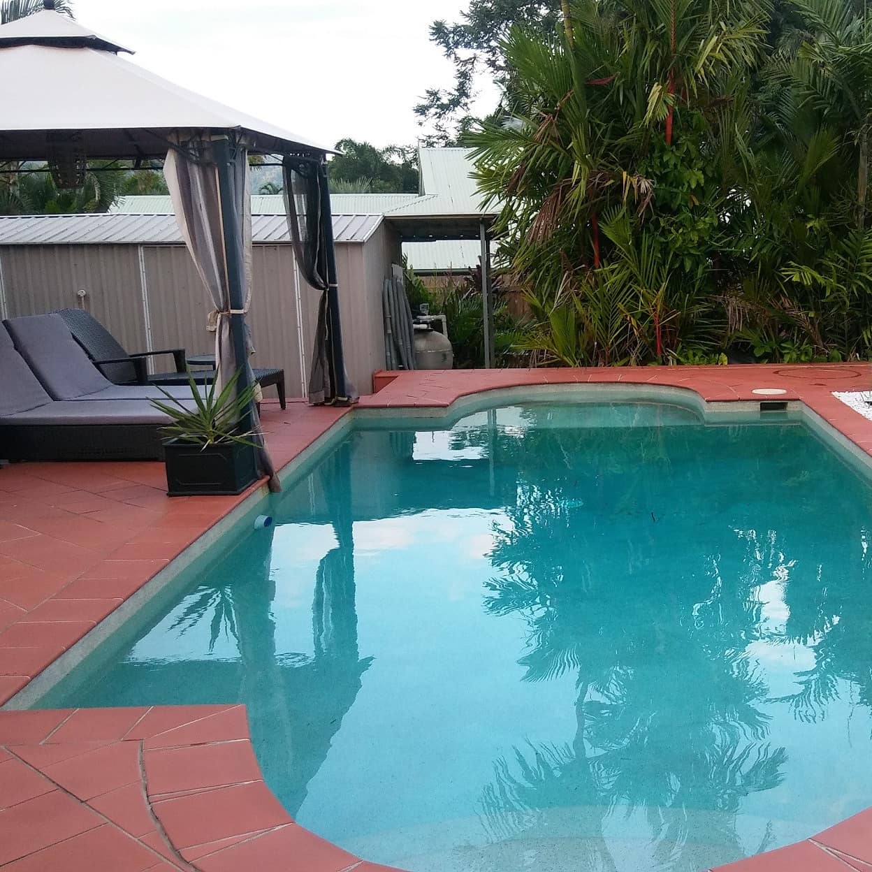 A pool and patio space, red tile, white gravel plant beds, cabana with grey lounge chairs, and lush tropical greenery.