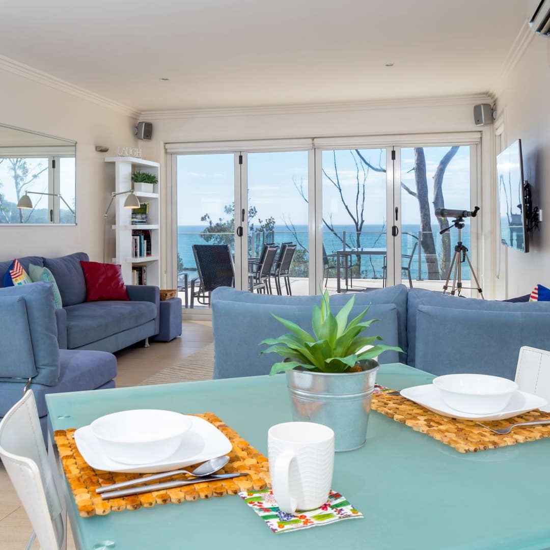 Bright open living and dining room of beachfront apartment with tile floors, light blue furniture and ocean view. 