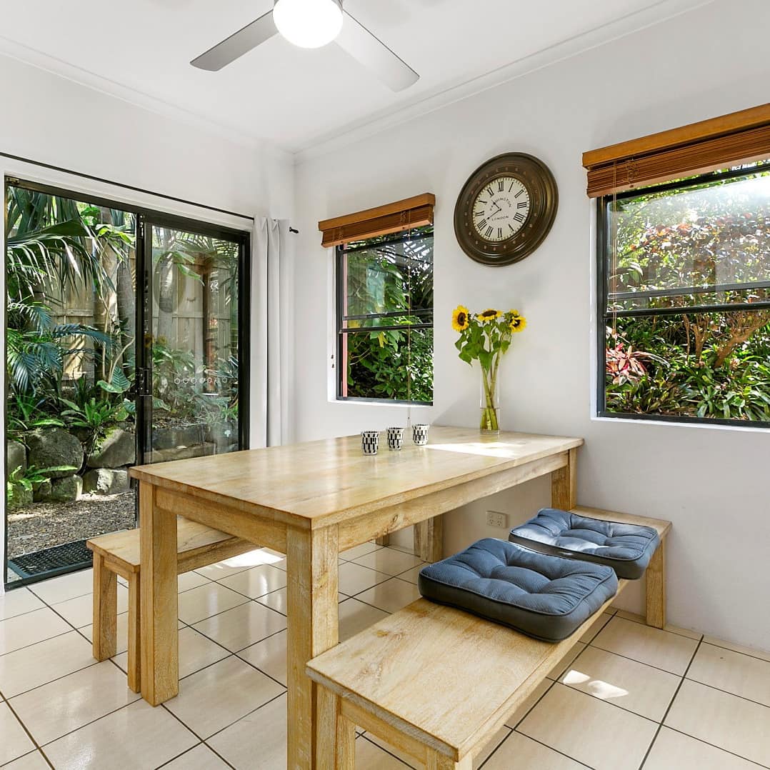 Sunny dining area with beige tile floors, white walls, wooden dining set, sliding glass doors to a lush yard.
