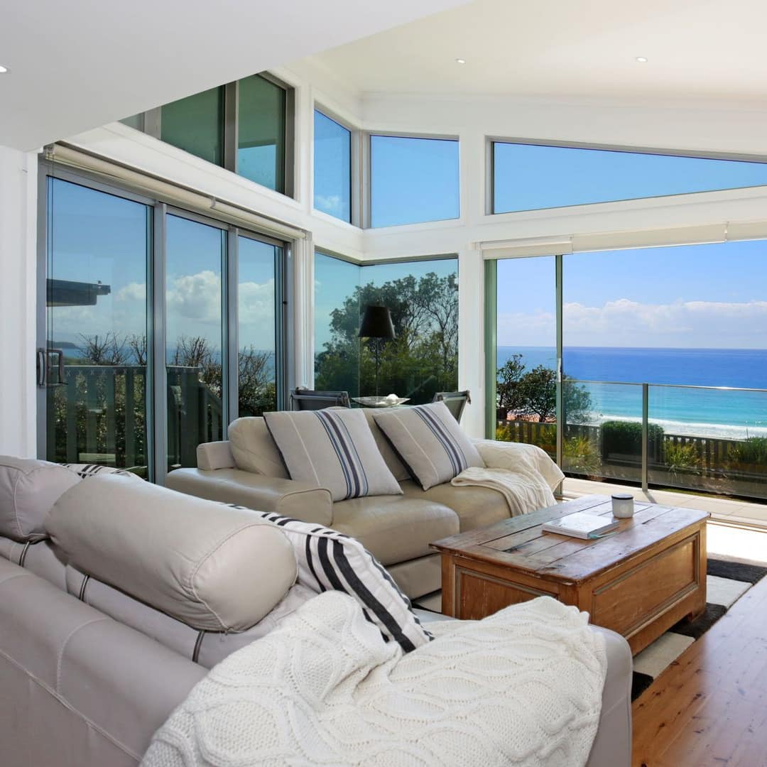 Open living room, floor to ceiling windows with ocean views, hardwood floors, beige leather sofas, and wood coffee table.
