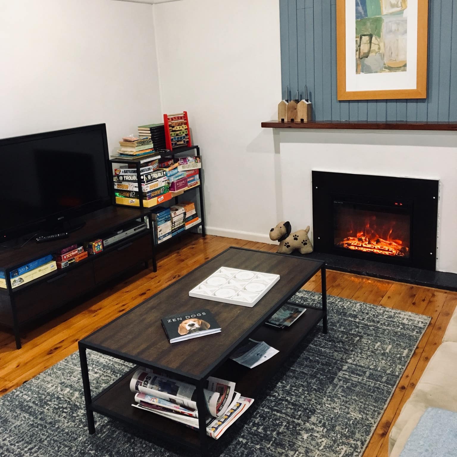A living room with hardwood floors, electric fireplace, and a television next to a bookshelf filled with puzzles and games.