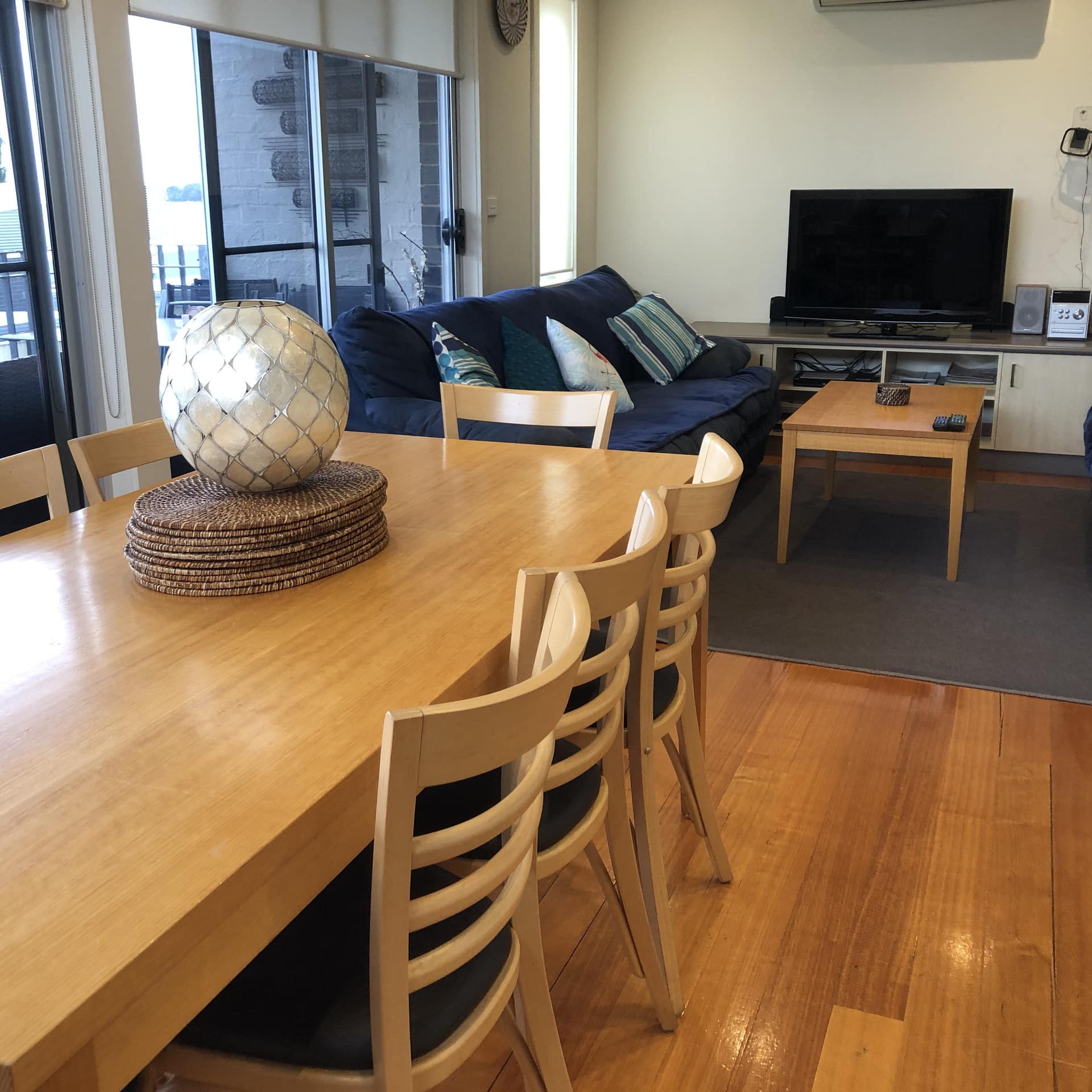 Light wooden table and chair aside a living area of couches and tv with side glass wall of windows and sliding doors