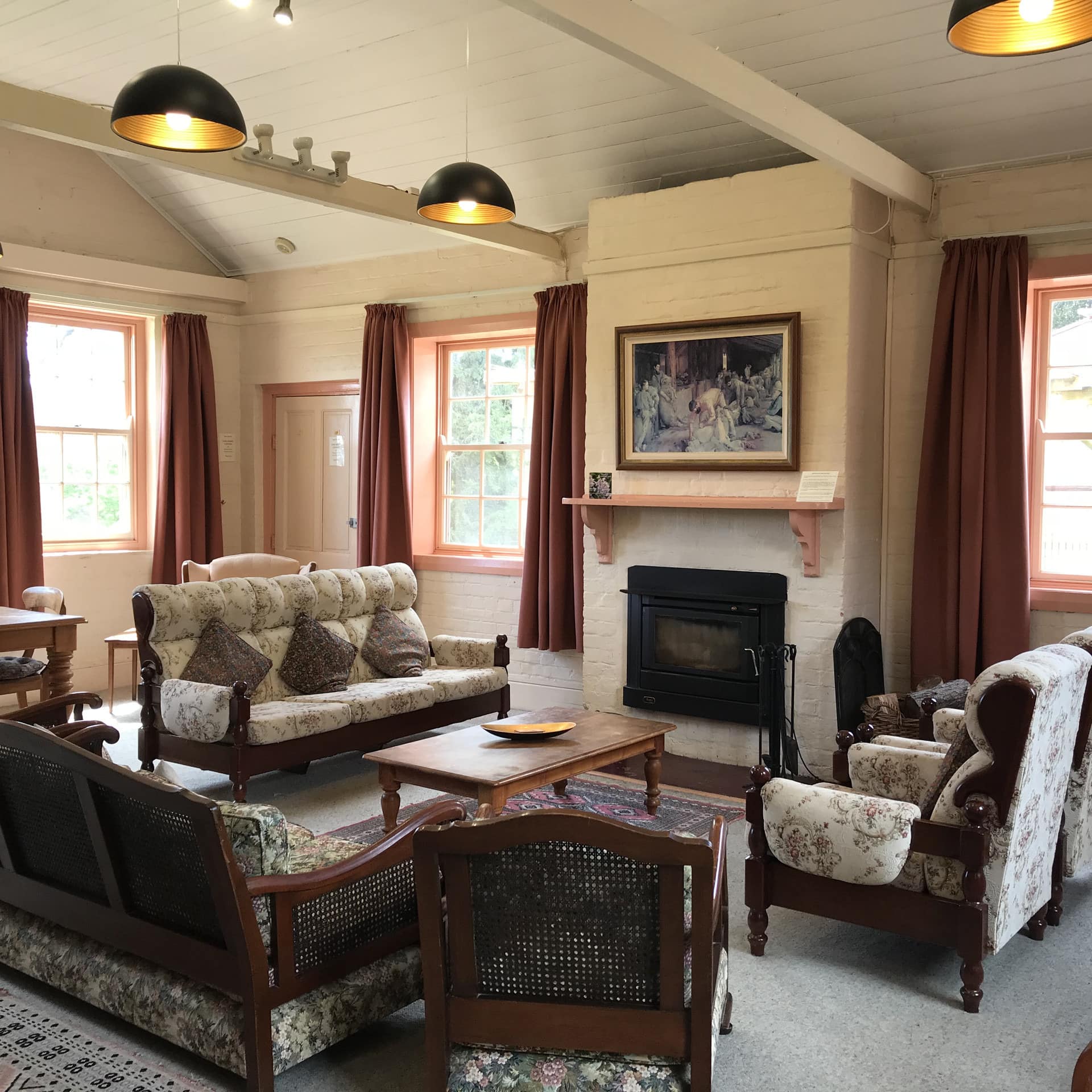 Open living room with white, brick walls, vaulted ceiling beams, fireplace, red window curtains, and antique furnishings.