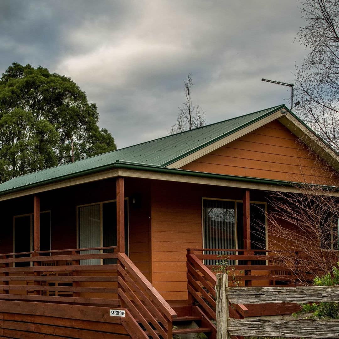 Wooden cabin with green roof, wrap-around porch, surrounded by trees.