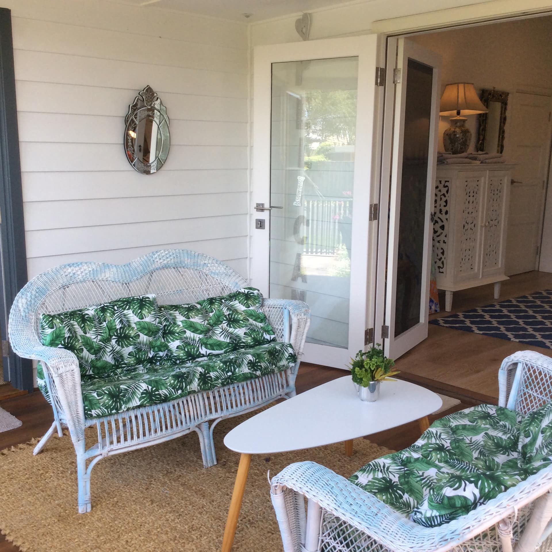 Wooden patio space with two white wicker sofas with palm tree print cushions, modern white coffee table, woven rug.