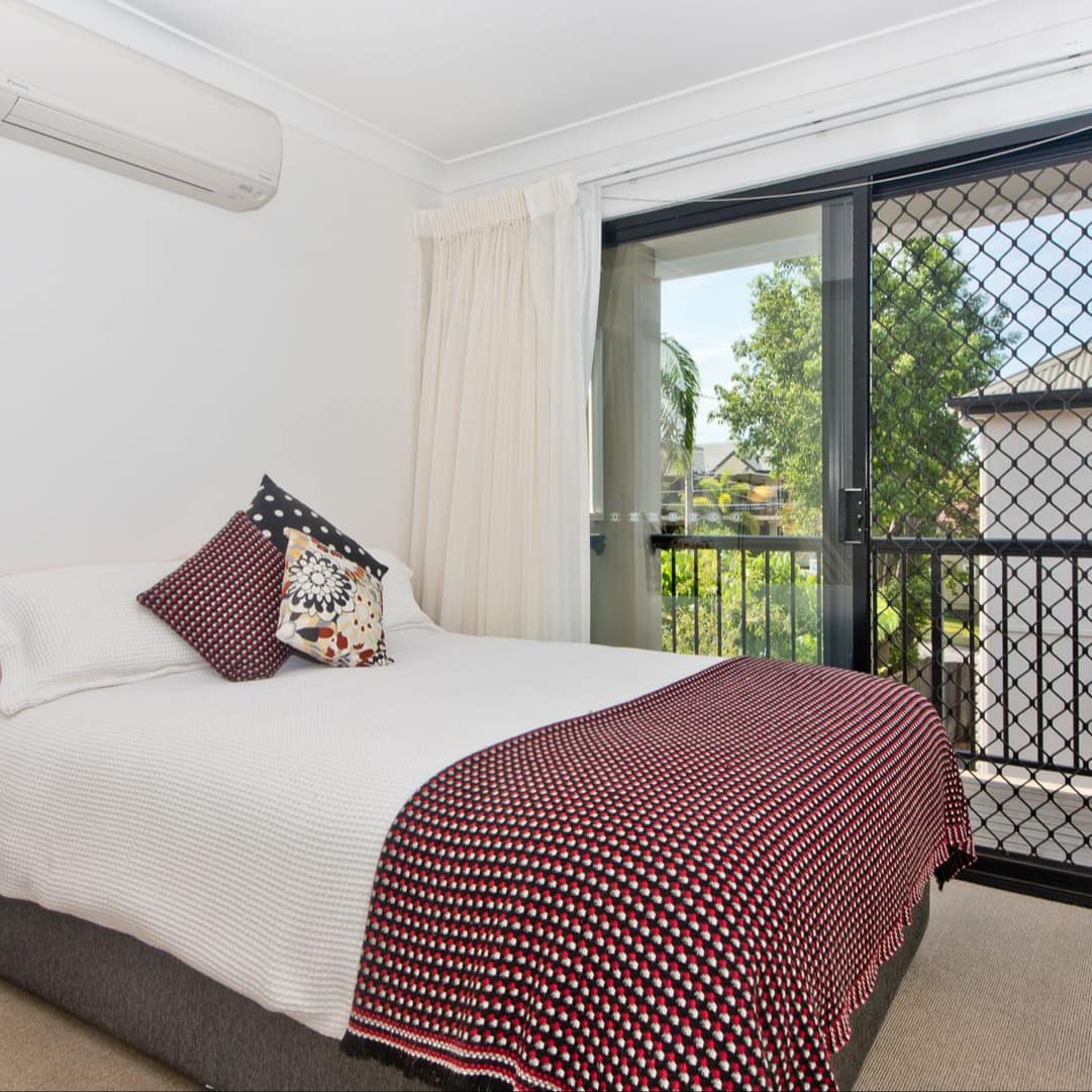 Bedroom with attached balcony, beige carpeted floors, white walls, a queen size bed with white, red, and black bedding.