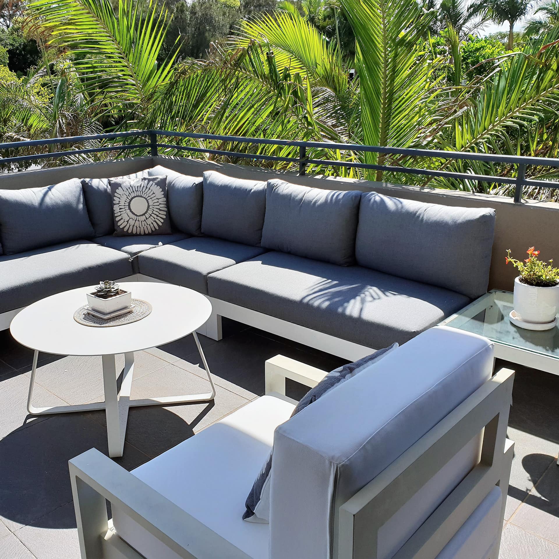 Grey tile patio surrounded by palm trees with a grey cushioned sectional, white chair and round, white table.