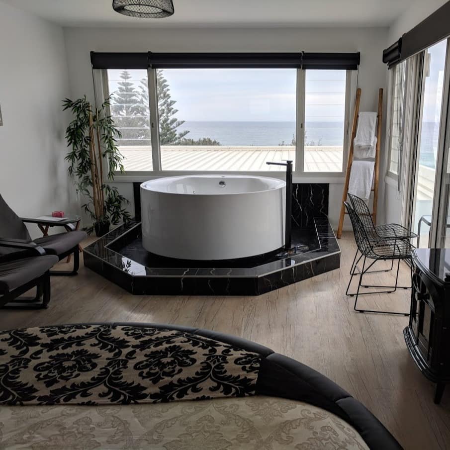 Studio apartment with hardwood floors, black and gold furnishings, a hot tub, and wide windows with ocean views.