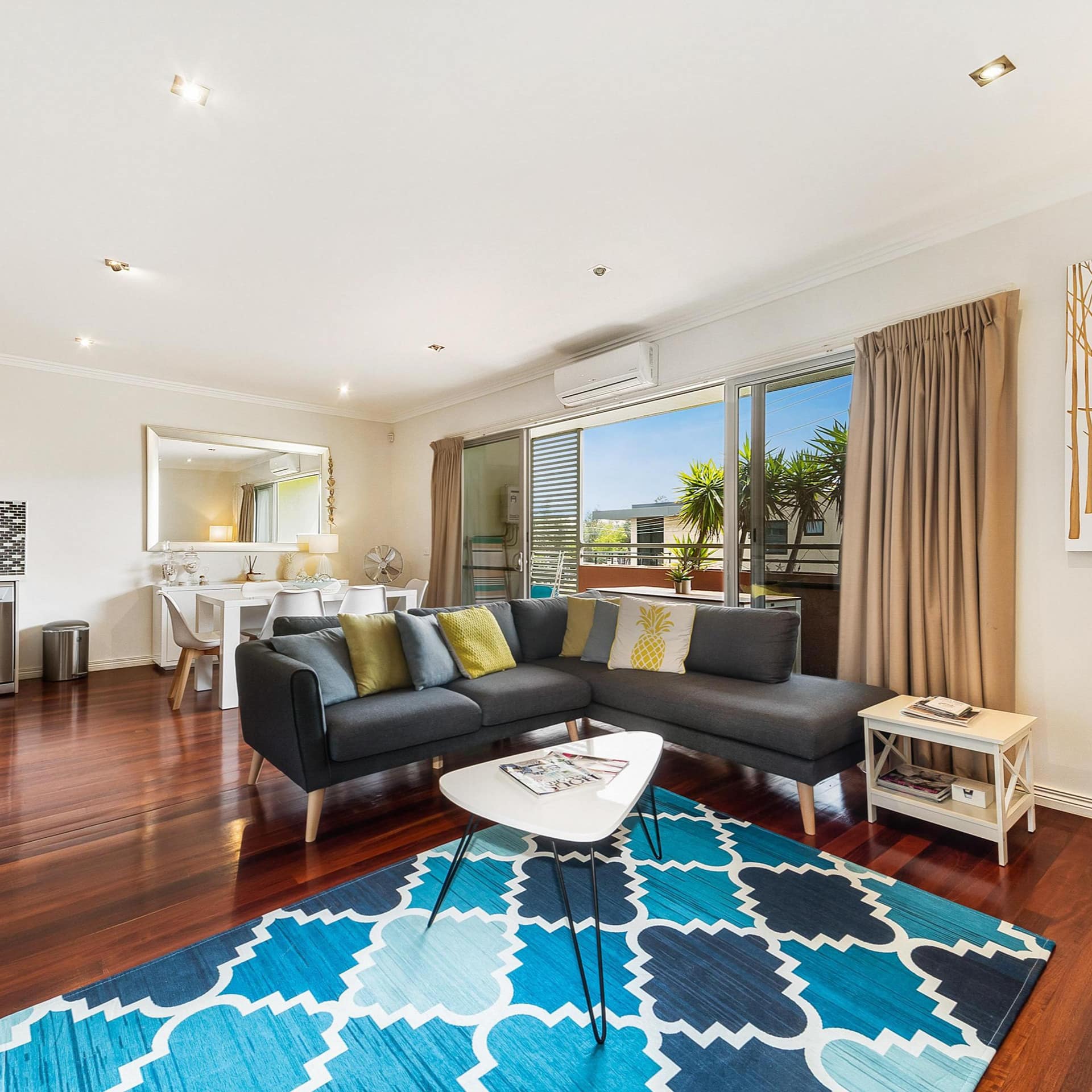 Open living room and kitchen with hardwood floors, grey sectional, blue and white rug, and wide windows to balcony.