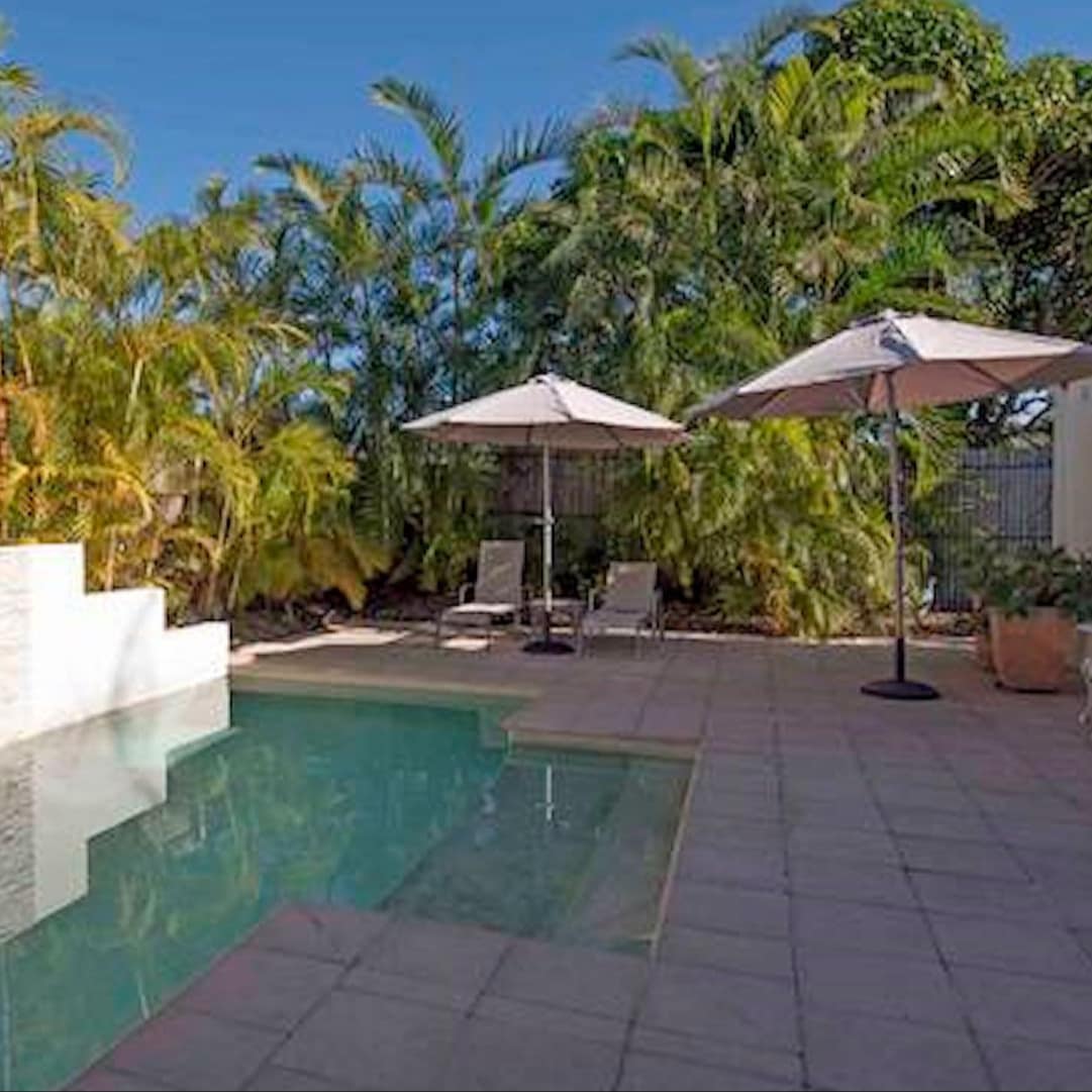 Private pool with grey tile pool deck, beige lounge chairs with umbrellas, surrounded by palm trees.