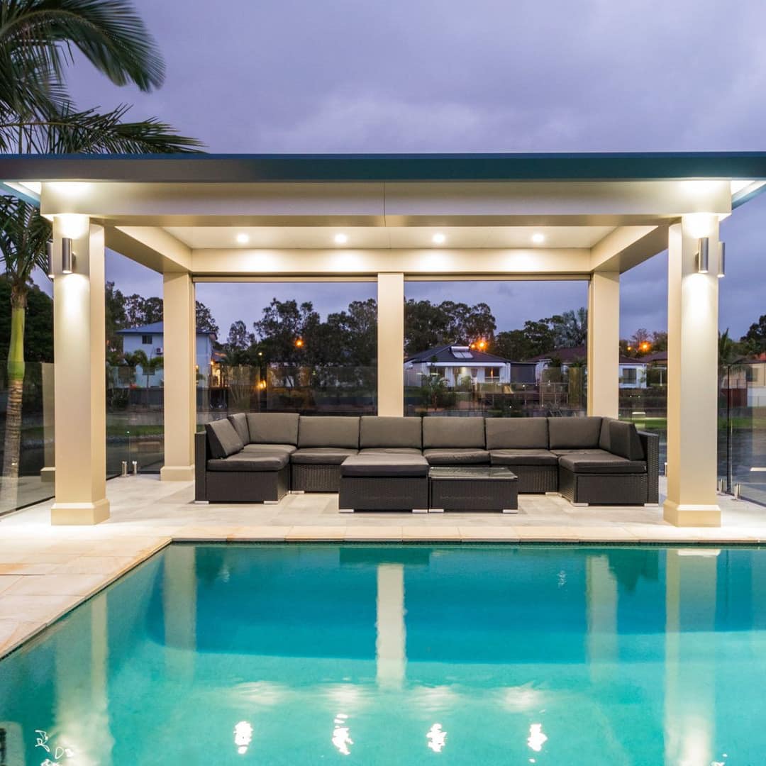 Nighttime view of an oceanside pool and pavilion, white tile floor, black outdoor sectional, and palm trees.