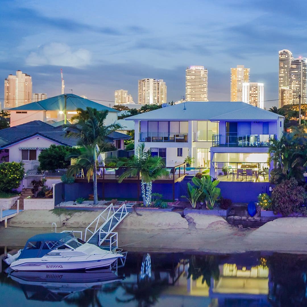 Evening view of waterfront luxury homes, a dock and boat in front of one home, brightly lit city skyline in the distance.