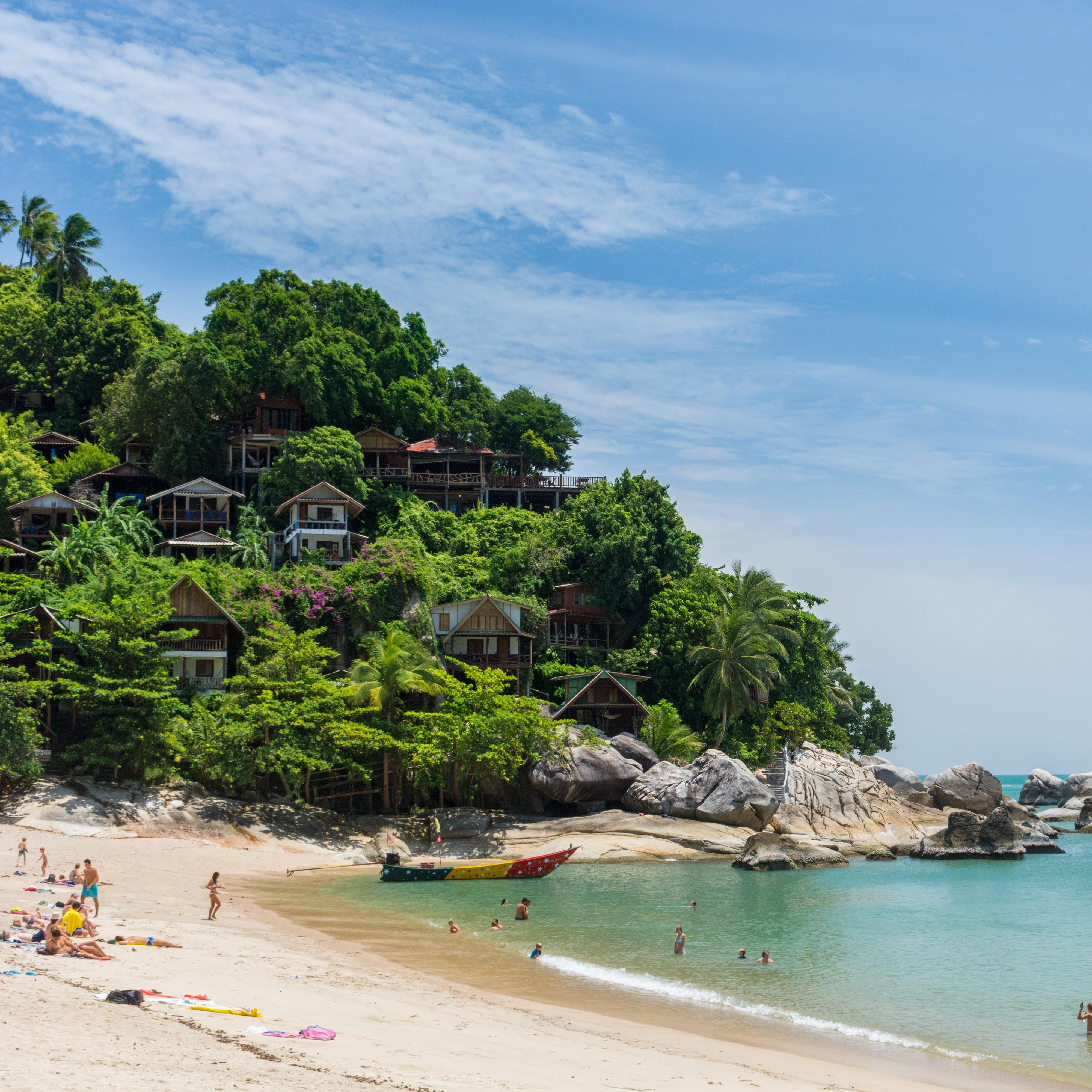People on the beach and in water with homes on the lush cliffs in Koh Phangan Thailand