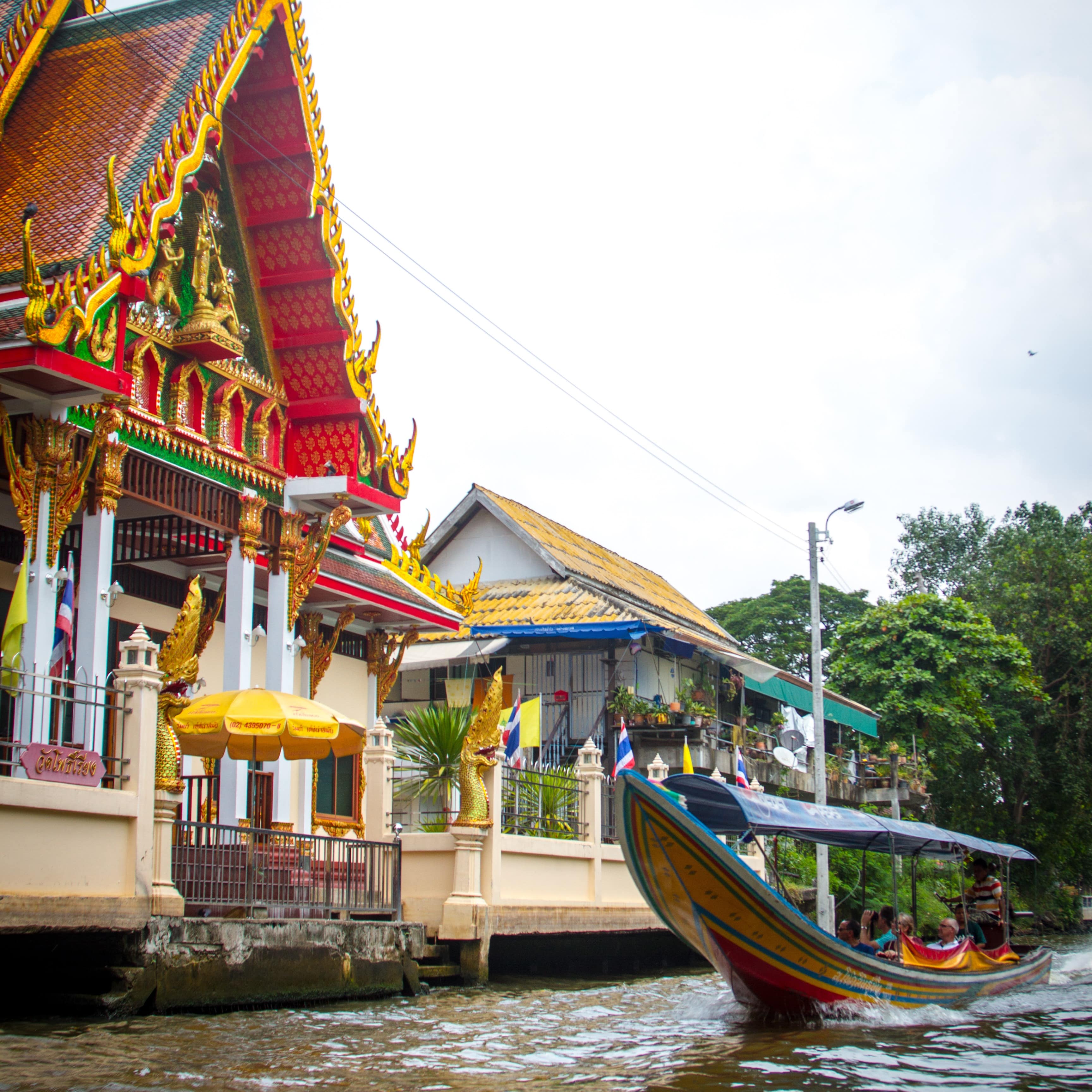 People in long tail boat motoring past a Buddhist temple in the canals in Bangkok, Thailand.