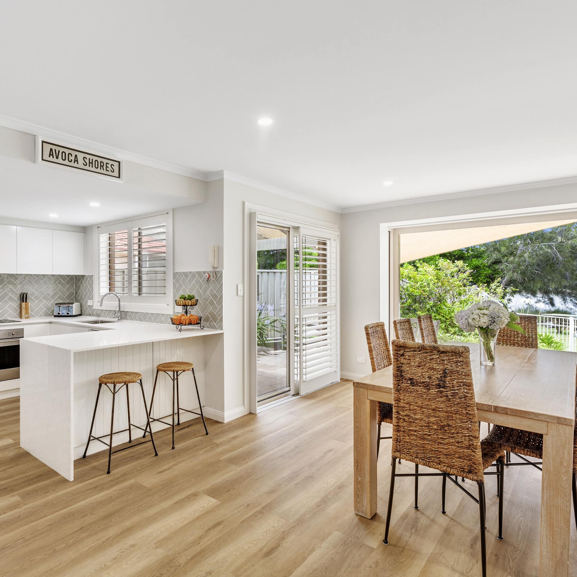 Open kitchen and dining room with folding glass doors to a garden, hardwood floors, wood dining table, wicker chairs.