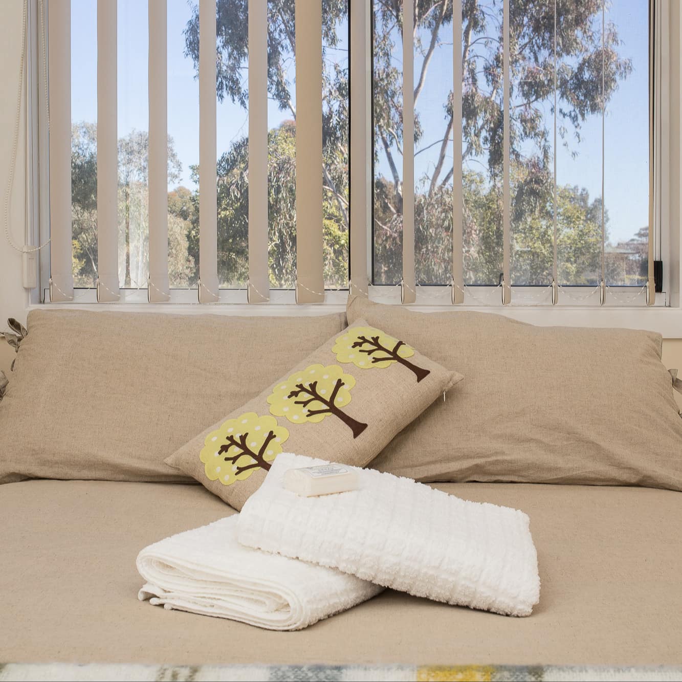 Beige bedroom with a queen size bed beneath a window with forest views, tan rustic bedding, and folded towels on the bed.