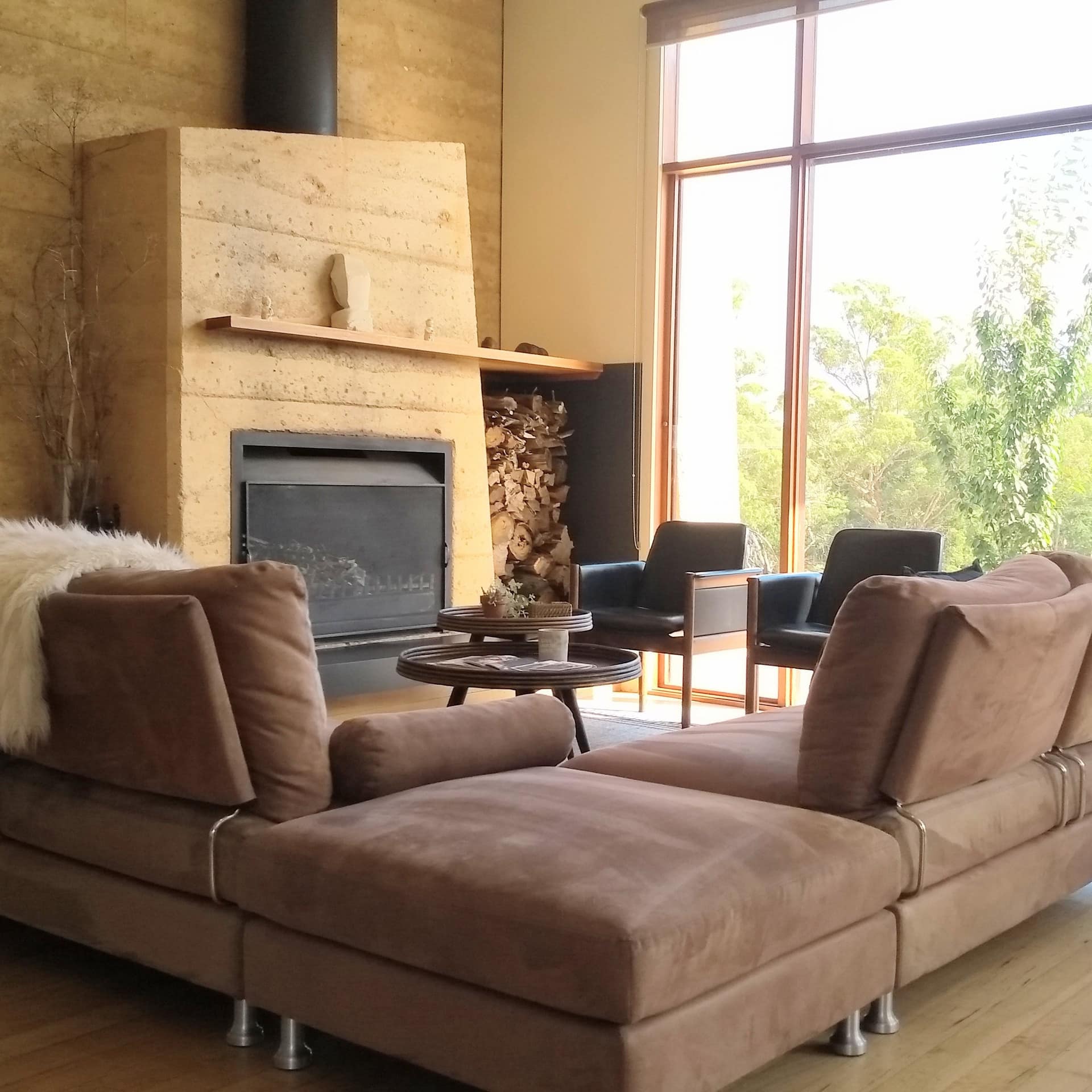A modern living room with wood-panelled walls, a brown suede sectional, fireplace, and floor to ceiling windows.