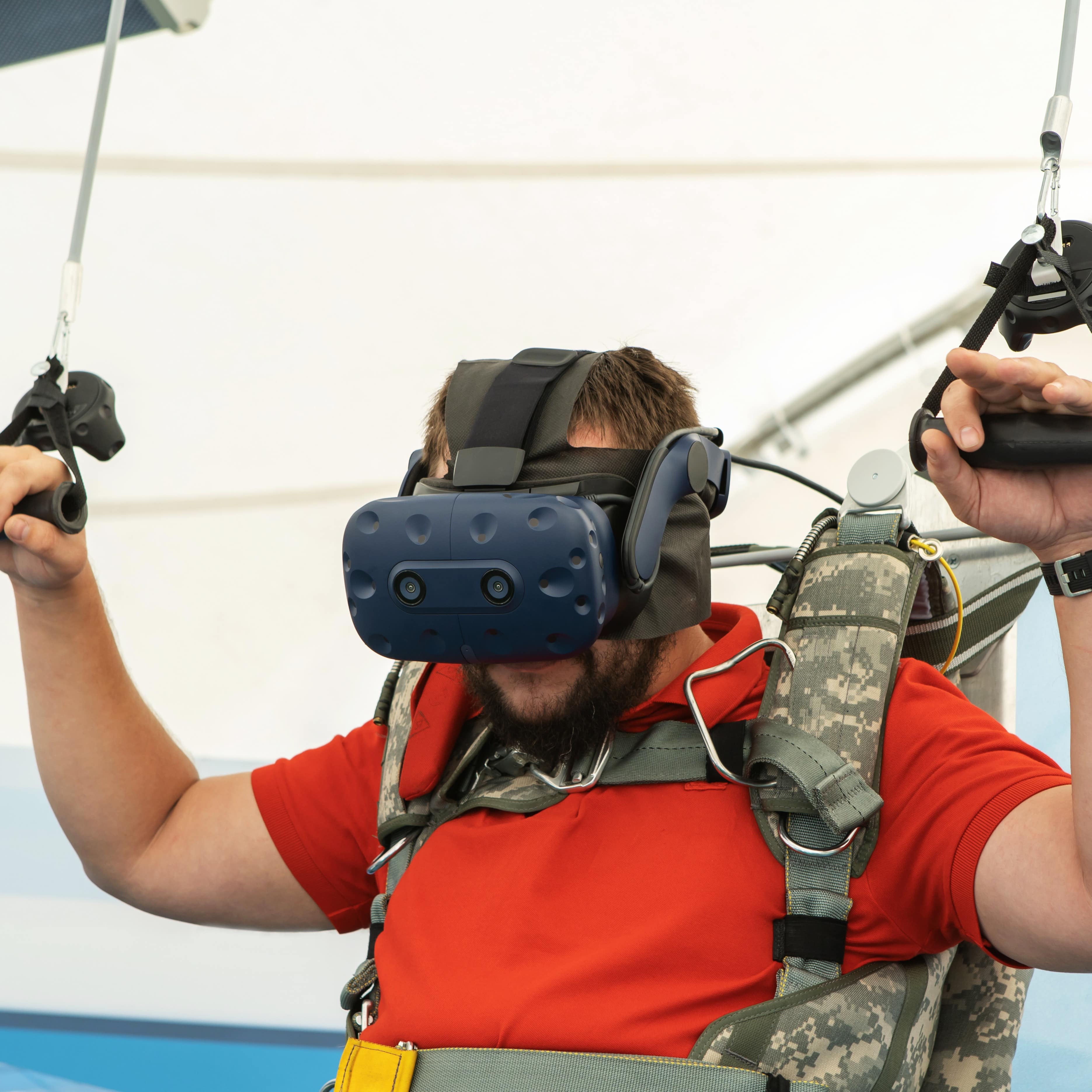 A man with red shirt wears virtual headset while experiencing a parachute jump on a 3D simulator