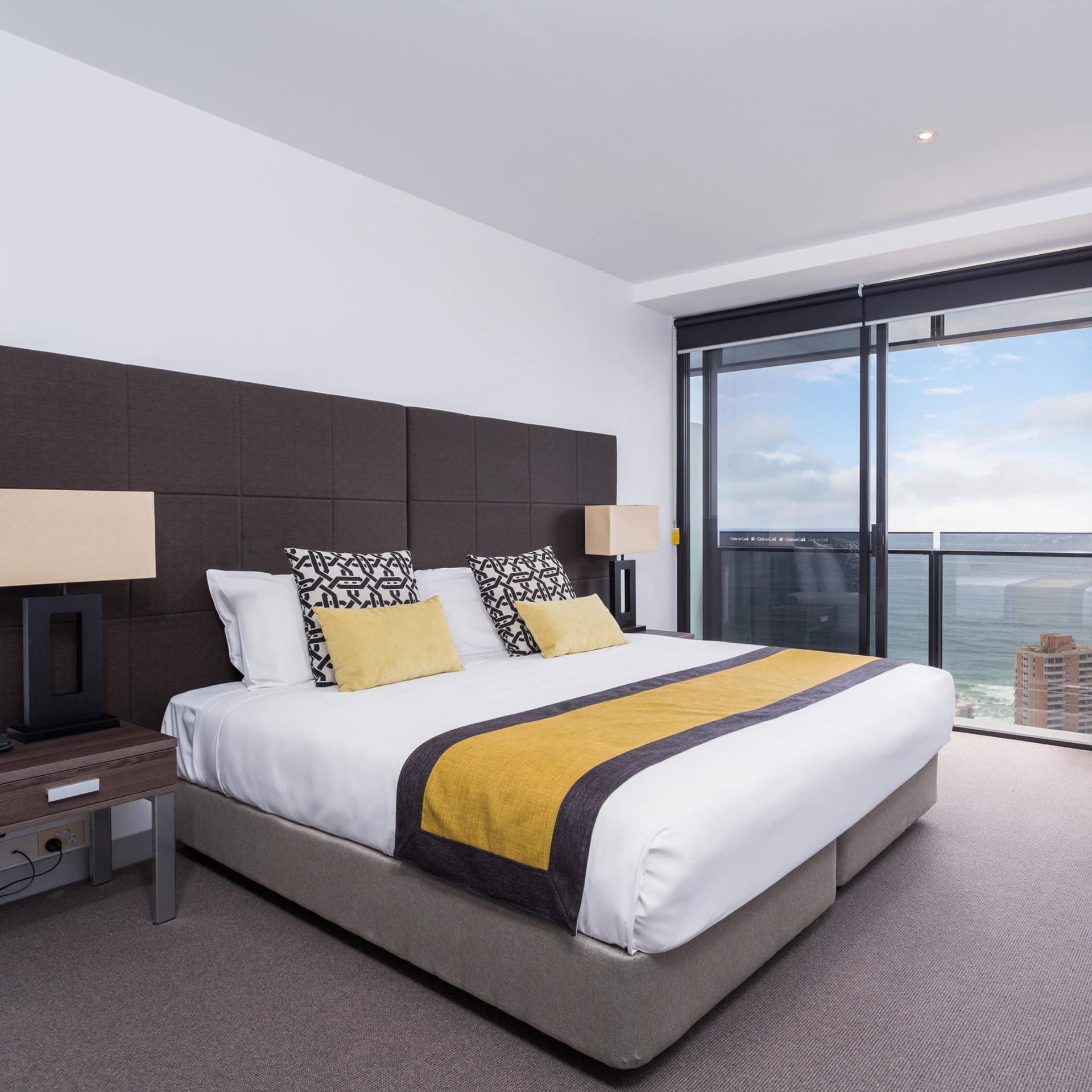 Bedroom with grey carpet, white walls, a king size bed with white and yellow bedding, and tall windows with city views.