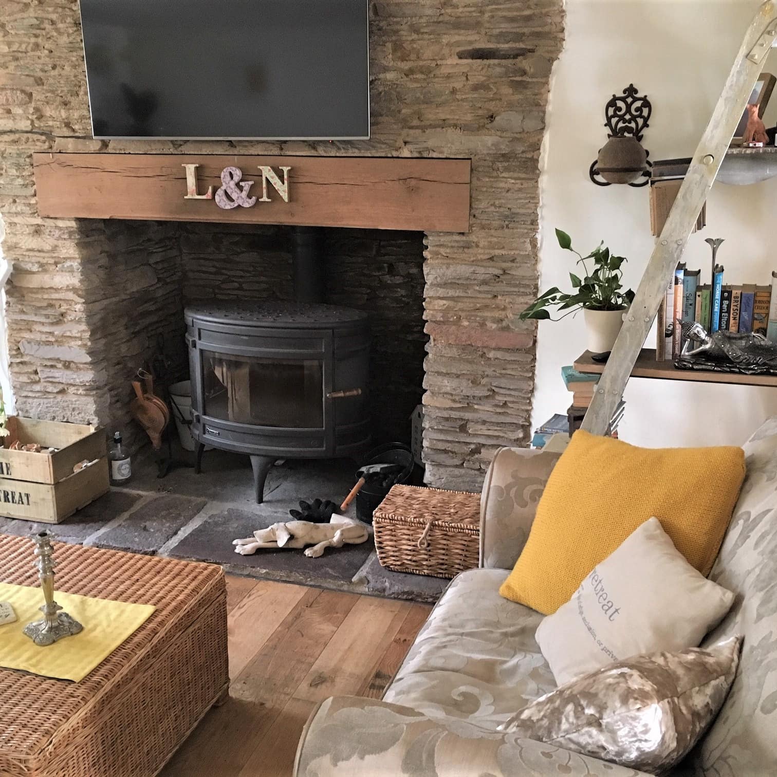 B&B living area with wood stove in stacked stone fireplace, wicker coffee table, overstuffed sofa and books on shelves