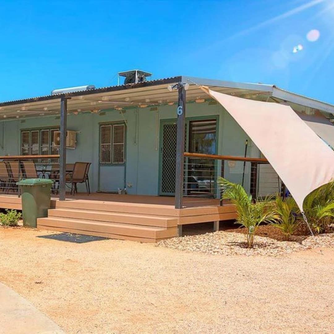 Exterior of a teal cottage, covered front porch with a dining set, pebble gravel driveway, plant beds with tropical plants.