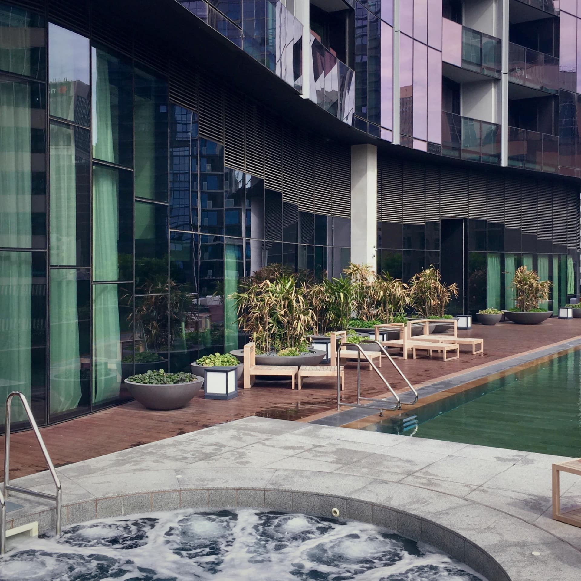 View of a pool and hot tub alongside a modern high-rise building with wooden pool deck and wooden lounge chairs.