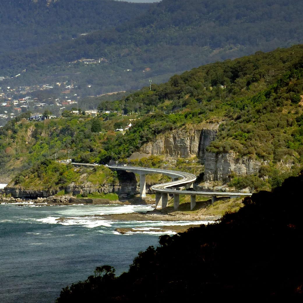 A view of Sea Cliff Bridge, a balanced cantilever bridge, by white cliffs and the sea with hills covered in trees and shrubs