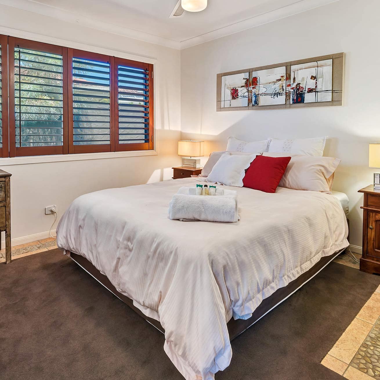 Bedroom with stone tile floors, a king size bed, white and red bedding, wide windows with wood slate blinds.