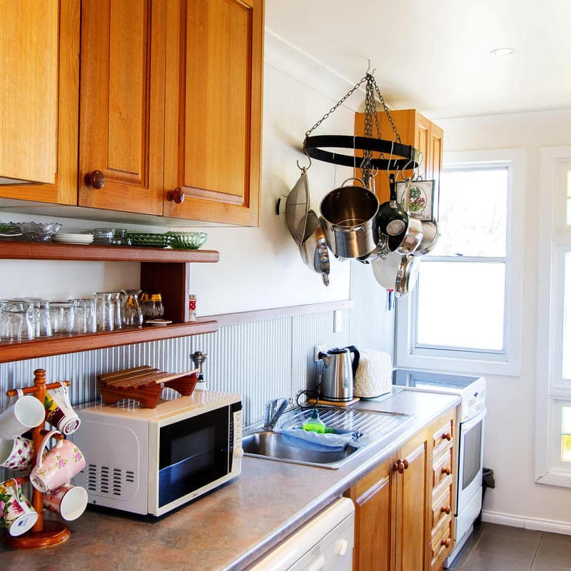 Galley kitchen with wood cabinets, metal backsplash, wood shelves with glass cups and teacups, and a stained glass window.