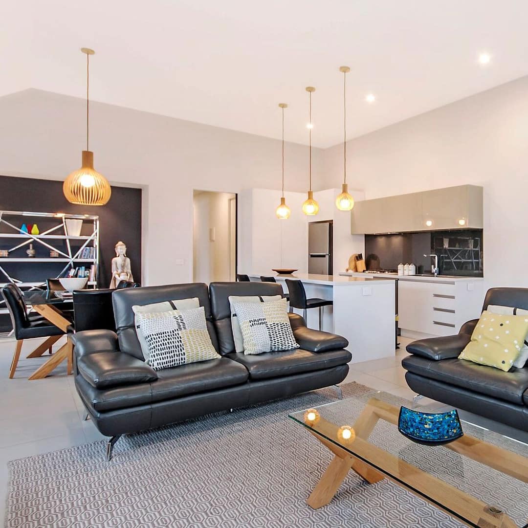 Modern, open living room and kitchen, black leather sofas, glass coffee table, white kitchen cabinets, black dining set.