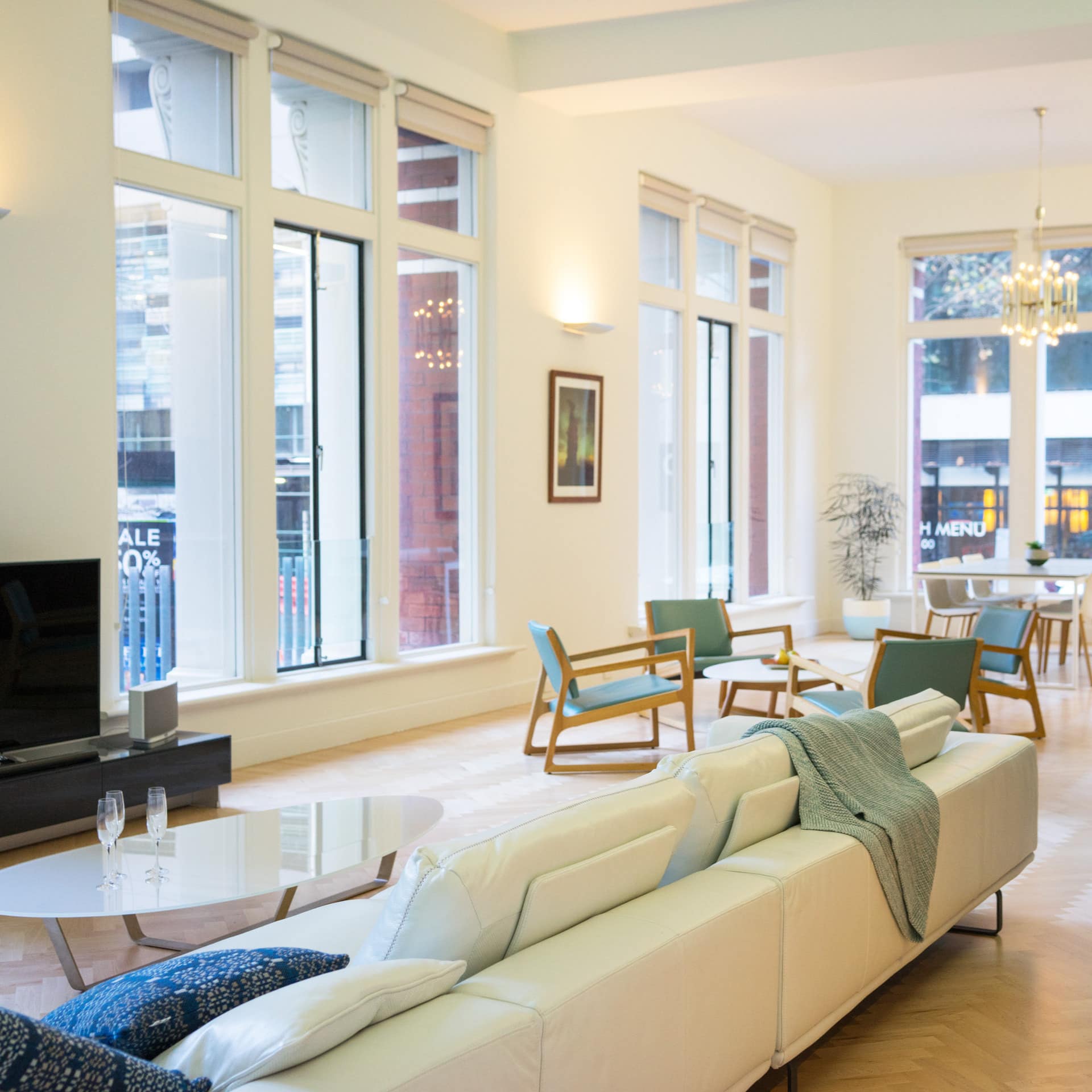 Open living and dining room with chevron wood floors, white leather sectional, flatscreen TV, and tall windows.