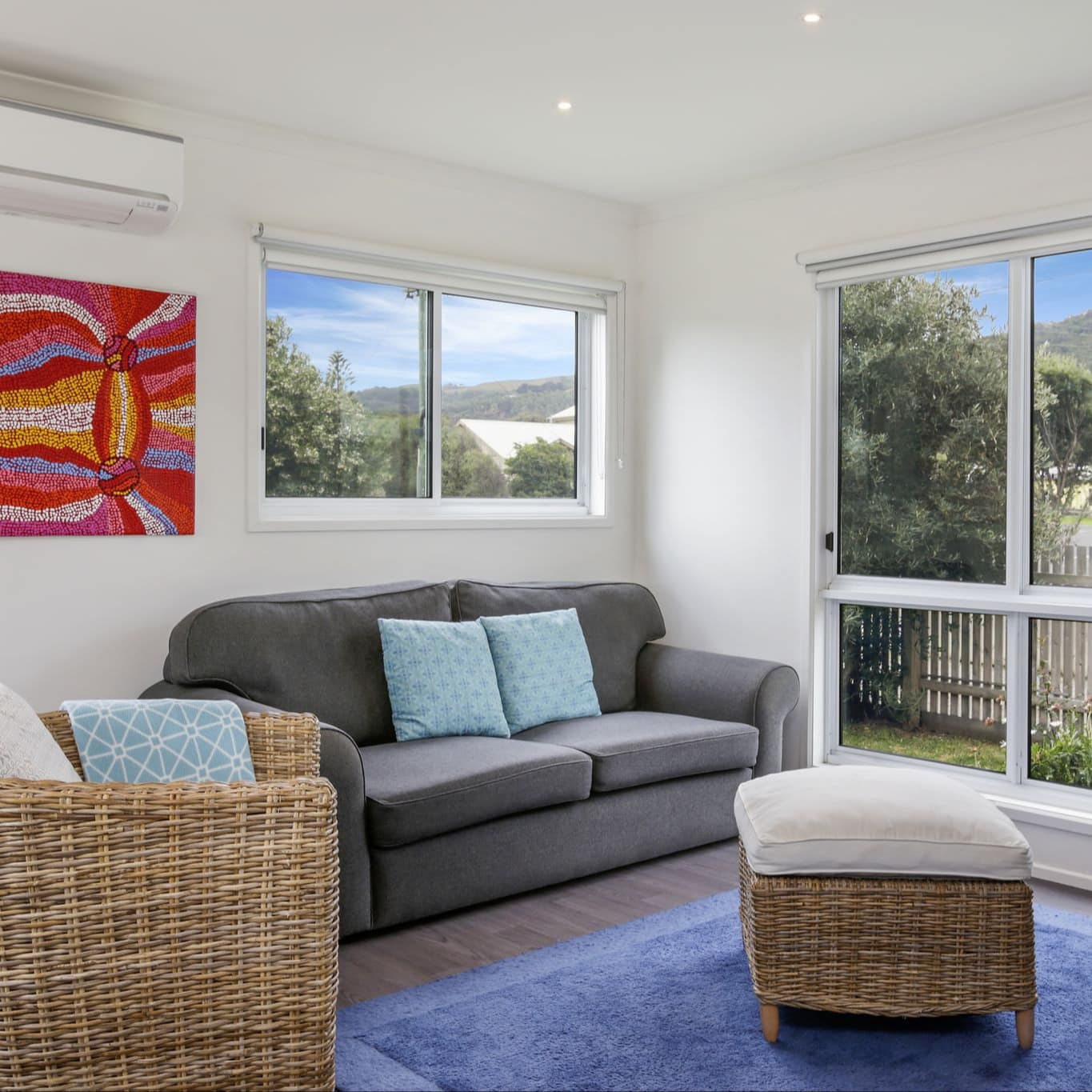 Living room with white walls, gray sofa, round wicker chair and foot rest, blue area rug, red abstract art and tall windows.