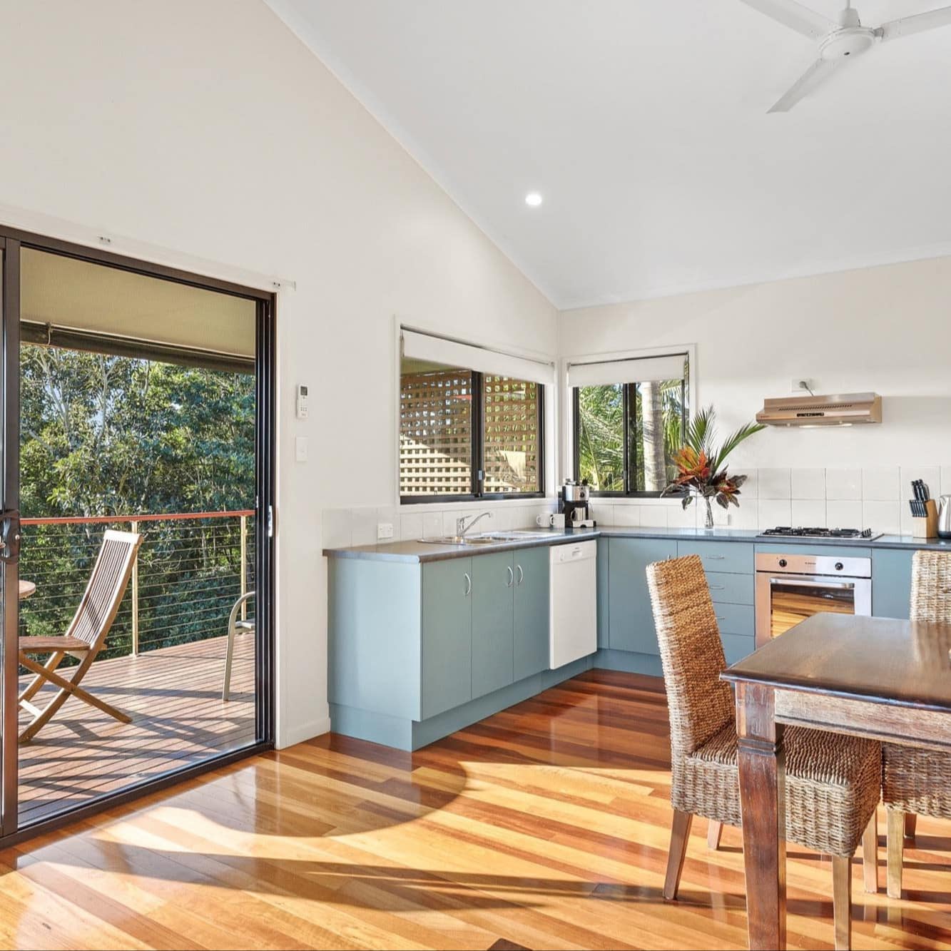 Sunny, open kitchen and dining room with attached veranda, blue kitchen cabinets, wood dining table and wicker chairs.