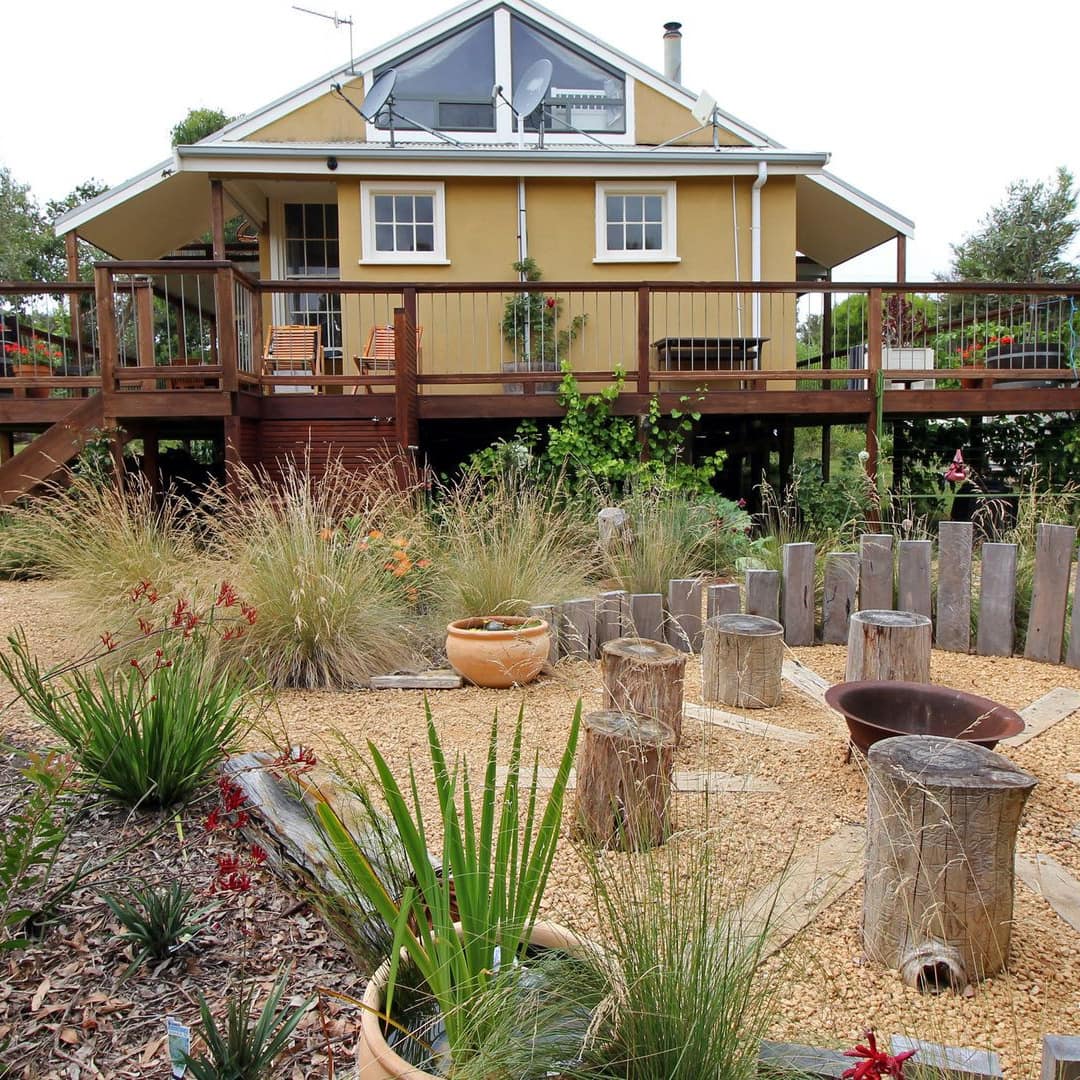 Exterior view of yellow cottage with wooden wraparound porch, landscaped lawn with plants and fire pit, tree-stump seats.